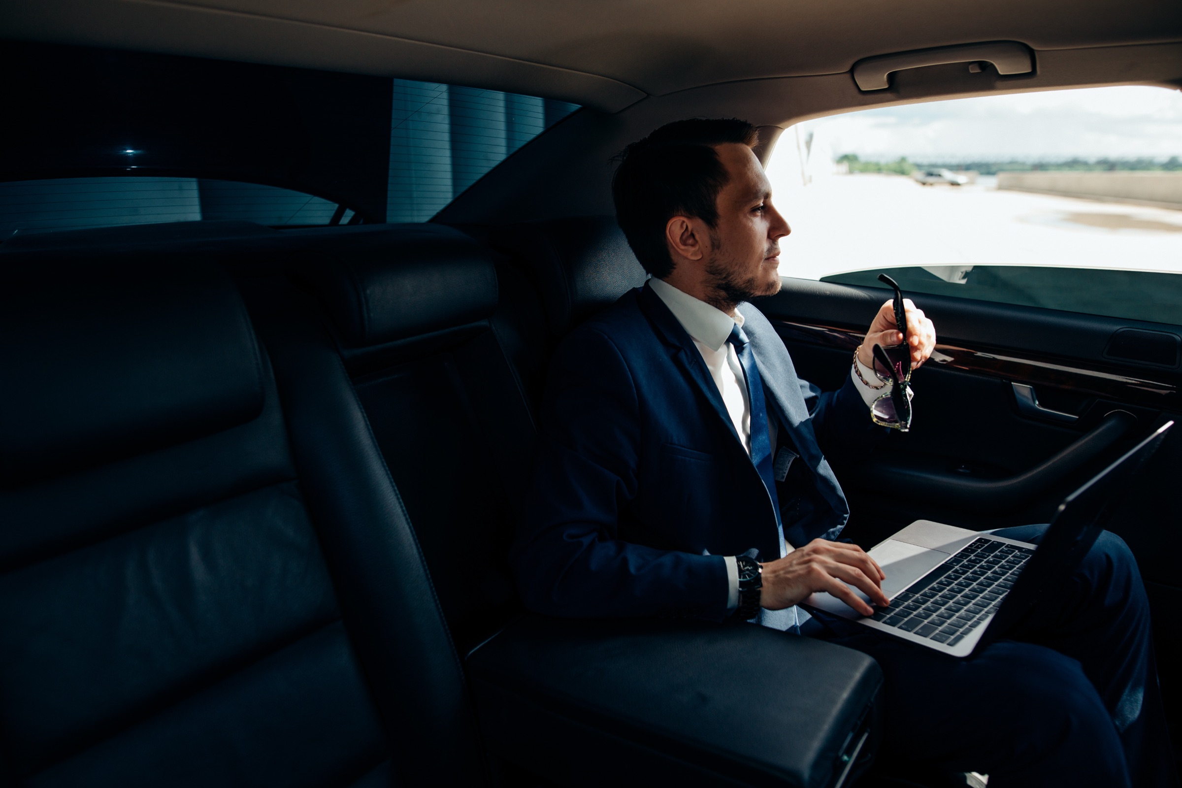 A passenger in a suit working on a laptop in the back seat of a car, looking out at an airfield.